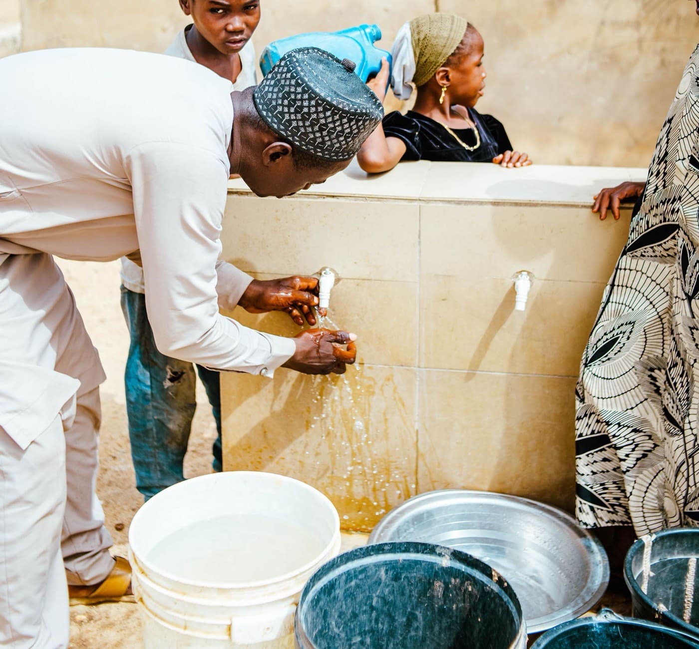 A man cupping water from a running tap at a tiled community standpipe, children waiting behind him with buckets